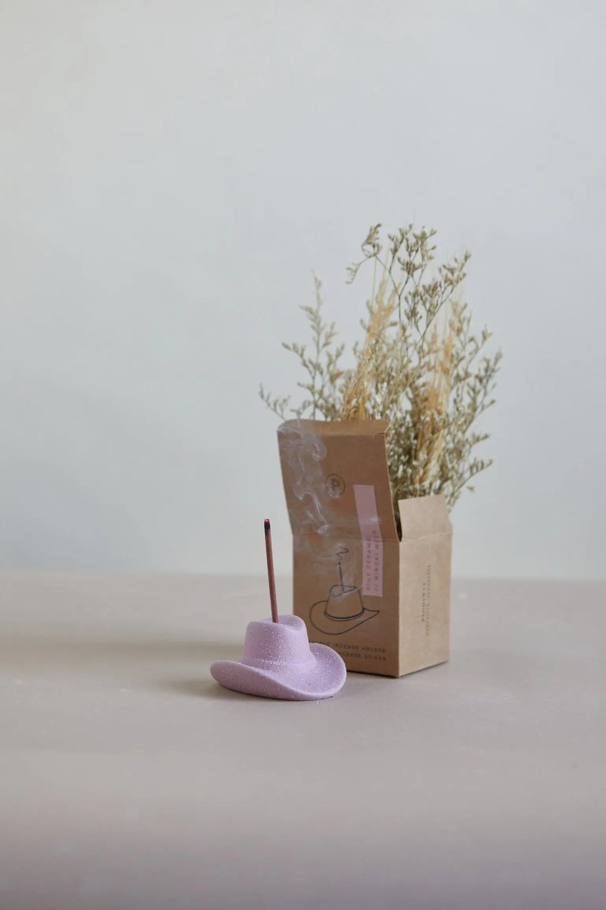 A Hinoki Wood - Incense Holder shaped like a small lavender cowboy hat holds a lit incense stick, placed in front of a brown paper bag with dried plants and set against a neutral background.