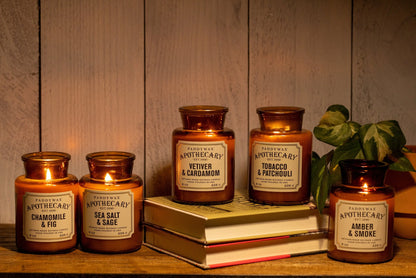 Five Amber + Smoke - Apothecary 8 oz Candles in brown glass jars are arranged on and beside two stacked books, with a small green plant in the background. Each candle features a luxury home fragrance label.
