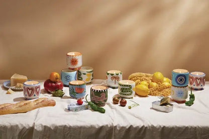 A white tablecloth holds a Lush Palms - A Dopo 8 oz Candle, surrounded by vegetables, lemons, pasta, cheese, a baguette, and small plates for a Mediterranean-inspired setting.