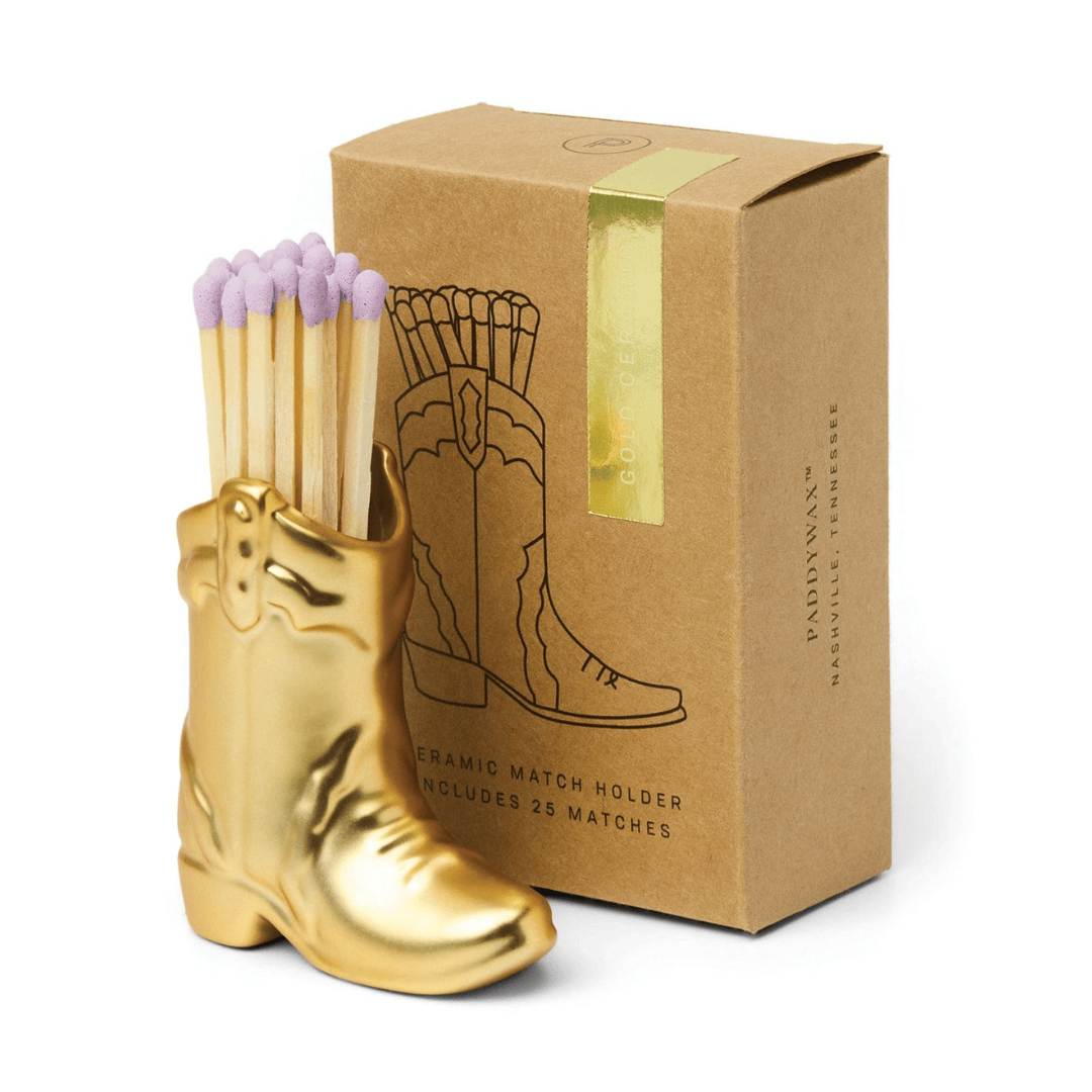 The Cowboy Boot Match Holder - Gold Metallic cradles lavender-tipped matches and sits beside a brown box adorned with a minimalist black boot illustration and product details—perfect as a unique Nashville gift or for adding Western charm to your home decor.