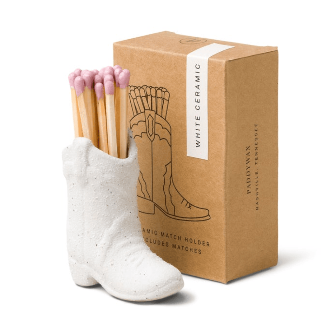 A Cowboy Boot Match Holder - White, filled with pink-tipped matches, sits beside a brown box labeled “White Ceramic” and decorated with a boot illustration—making it a charming piece of Western home decor and the perfect Nashville gift.