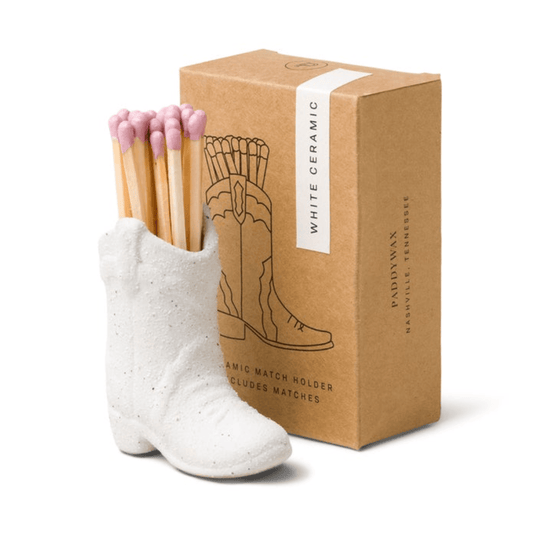 A Cowboy Boot Match Holder - White, filled with pink-tipped matches, sits beside a brown box labeled “White Ceramic” and decorated with a boot illustration—making it a charming piece of Western home decor and the perfect Nashville gift.