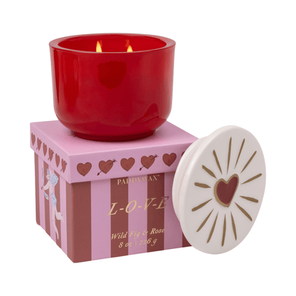 A Wild Fig & Rose - Charmed 8oz Candle is displayed on a pink and purple striped box labeled "LOVE" with hearts. Its white ceramic lid, adorned with a red heart and gold rays, rests beside the box.