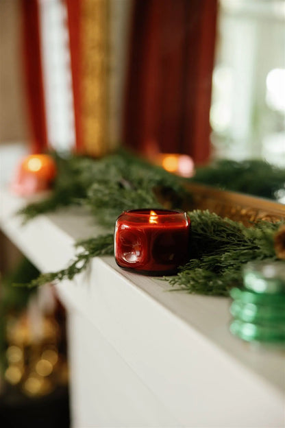 A red candle in a red glass vessel sits atop a white mantel decorated with evergreen branches; blurred green and orange candle holders, including the Yuzu Woods - Basalt 5 oz Candle, are also visible in the background.