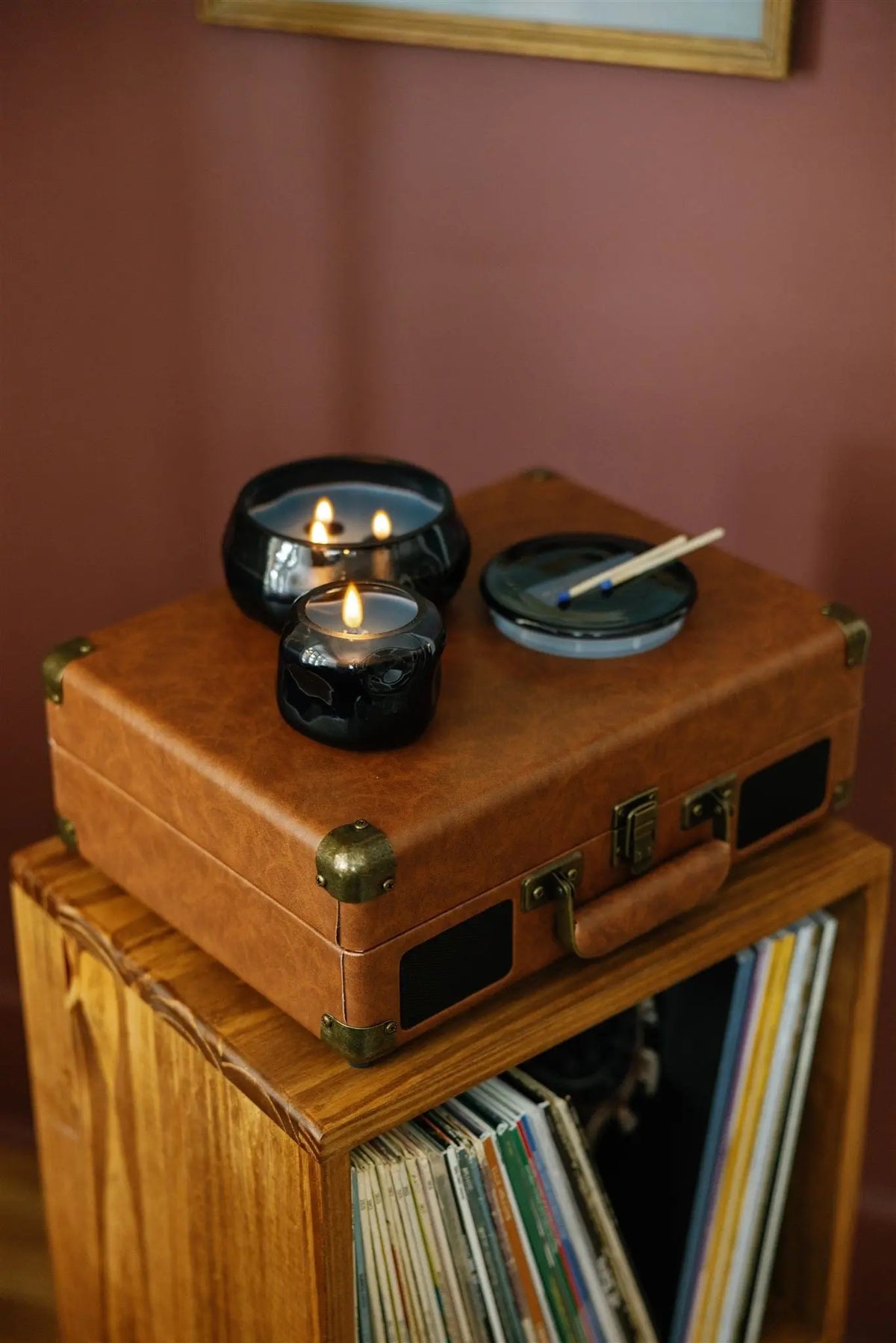 A lit Rosewater Santal - Basalt 10 oz Candle in a blue glass jar, alongside another candle and a dish with incense sticks, sits on a brown leather suitcase placed atop a wooden record stand filled with vinyl records against a reddish-brown wall.