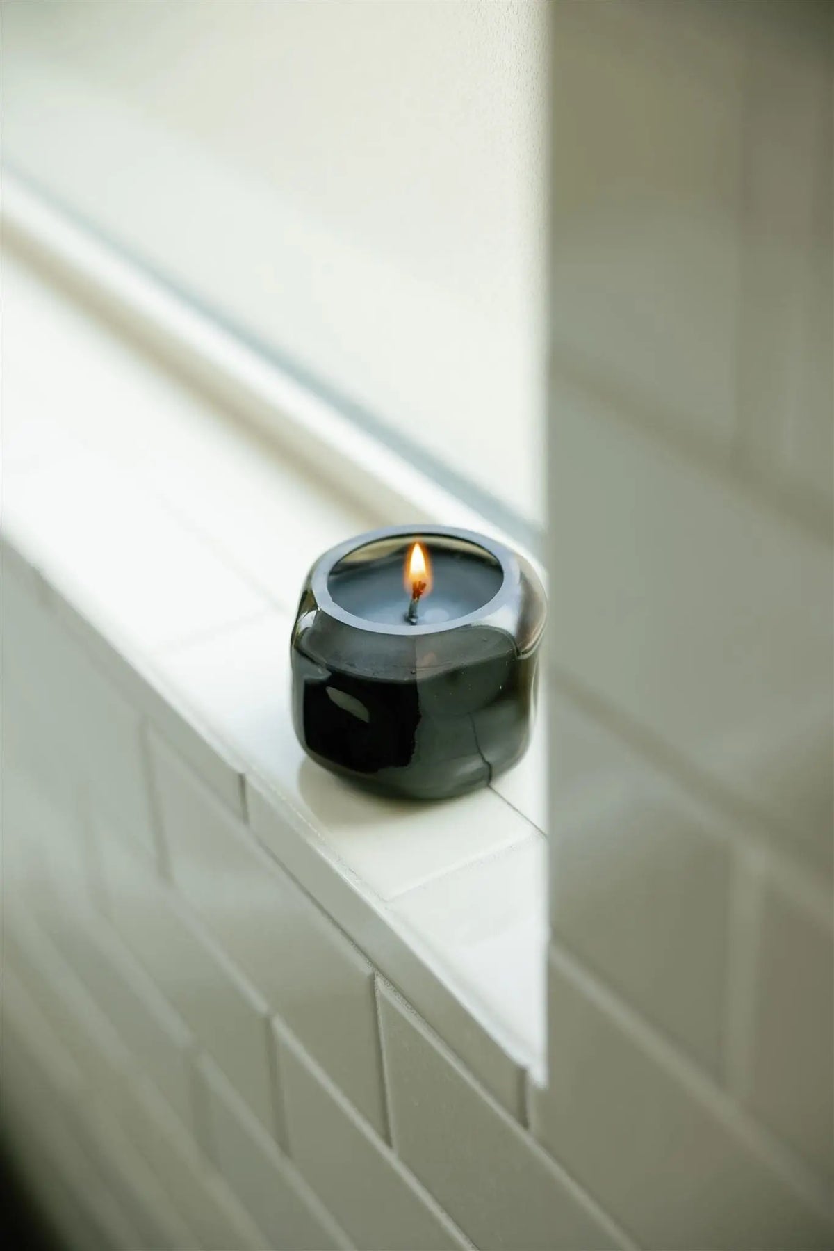 A Basalt 5 oz Palo Santo Suede candle glows on a white tiled windowsill, filling the calm, minimalist space with its subtle smoky wood fragrance.