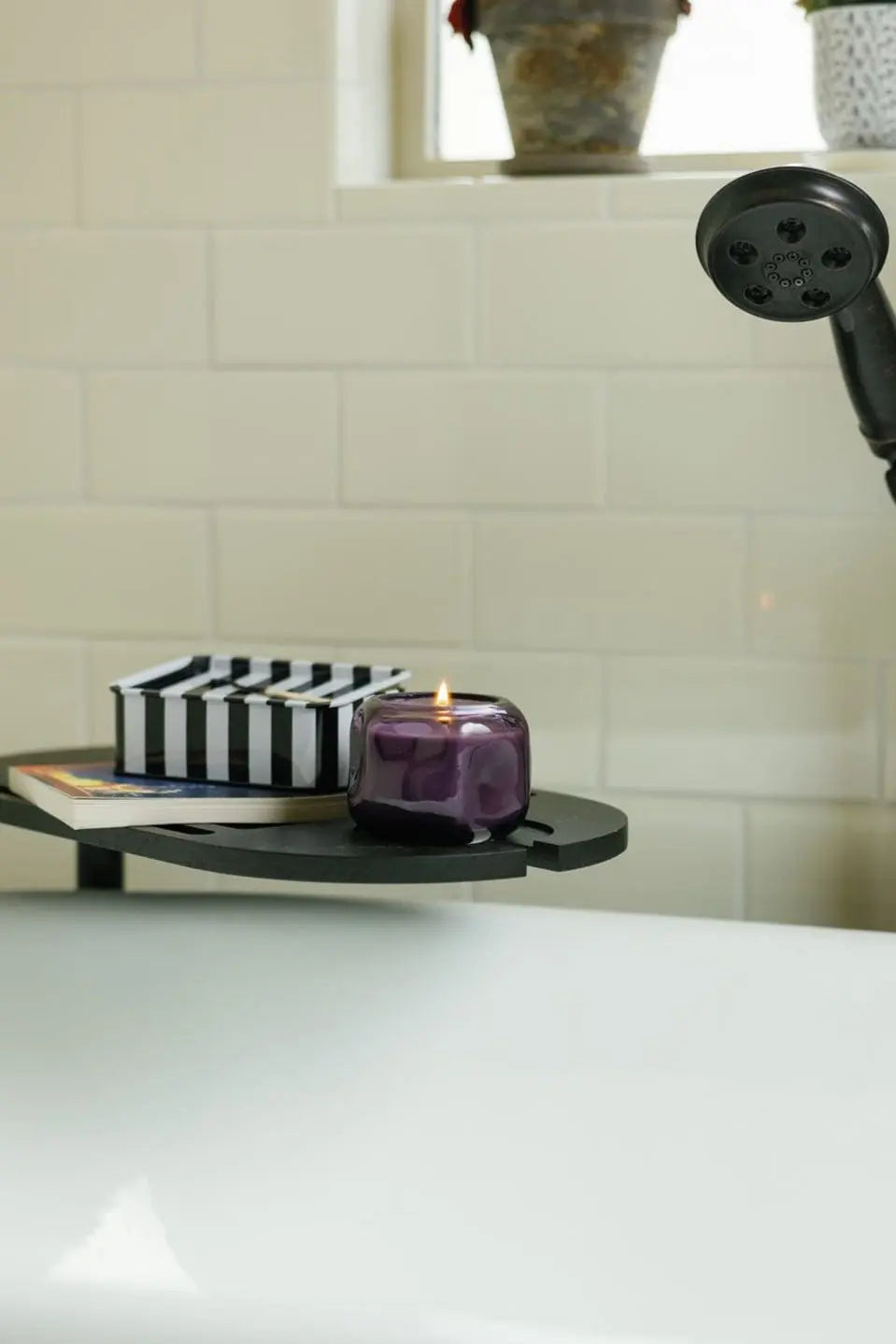 A Violet & Vanilla - Basalt 5 oz Candle in its elegant purple glass sits lit on a tray over a white bathtub, accompanied by a black-and-white striped box. The scene is set against a backdrop of white tiled walls with glimpses of a plant pot and showerhead.