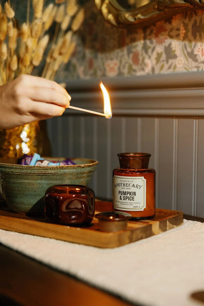 A hand lights a match above a brown glass candle labeled Sienna Sunset - Basalt 5 oz Candle on a wooden tray, with a ceramic bowl and a small brown cup nearby. The warm glow evokes an amber fragrance, while dried flowers and patterned wallpaper create an inviting backdrop.