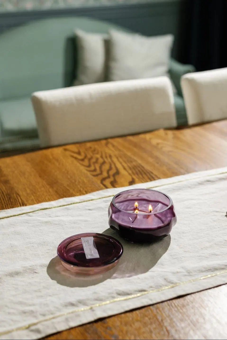 A Violet & Vanilla - Basalt 10 oz Candle, housed in an amethyst glass jar with two wicks, sits on a beige table runner atop a wooden table. The candle’s lid rests nearby, while white chairs and a couch with pillows are visible in the background.