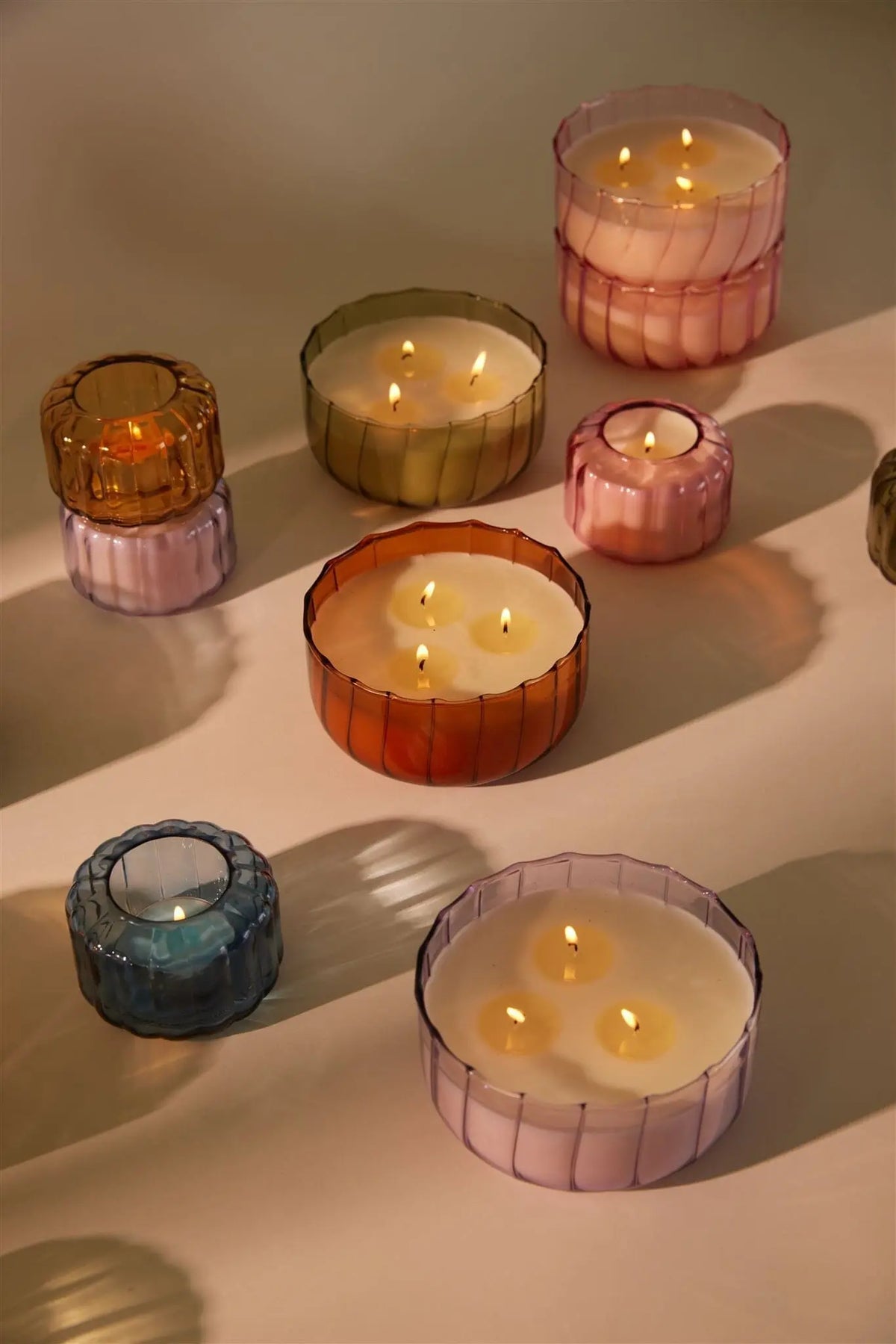A 12oz Desert Peach - 12 oz Candle and other vintage colored glass candles are displayed in colorful holders on a light surface, casting soft shadows.