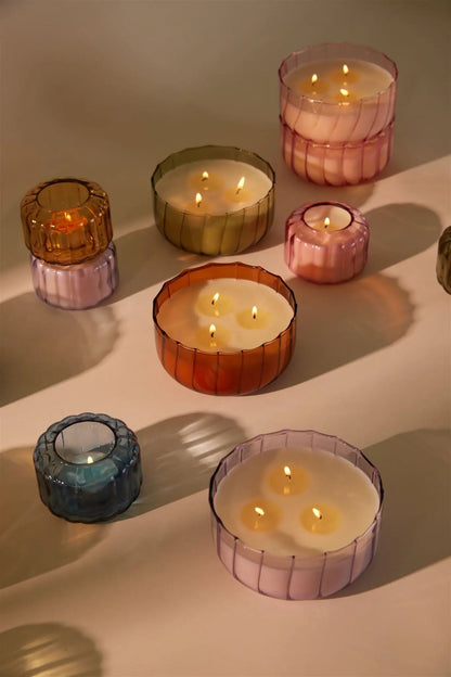 A 12oz Desert Peach - 12 oz Candle and other vintage colored glass candles are displayed in colorful holders on a light surface, casting soft shadows.