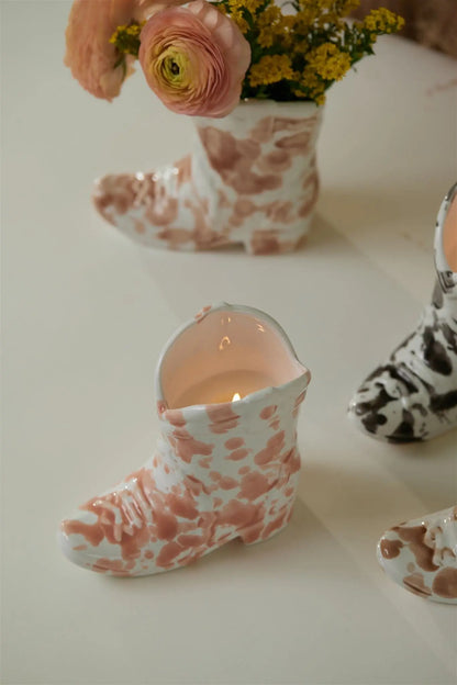 A Cactus Flower - Nashville Boot 6oz Candle burns inside a ceramic holder with pink and white spots. In the background, a matching boot-shaped vase holds flowers beside another boot with black and white spots.
