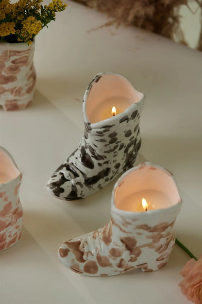 Two ceramic cowboy boot candle holders with brown and black spots hold lit candles on a white surface. A yellow flower arrangement and the Palo Santo Suede - Nashville Boot 6 oz Candle are partially visible in the background.