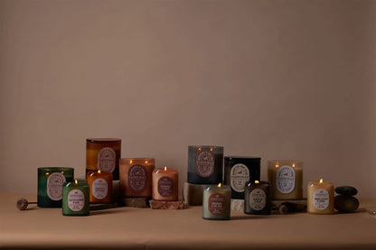The Vista 12 oz Candle in Rosemary + Sea Salt, with its vintage-inspired glass jar, is displayed on a beige surface with stones and wood accents, creating a cozy scene that evokes its warm, amber fragrance.