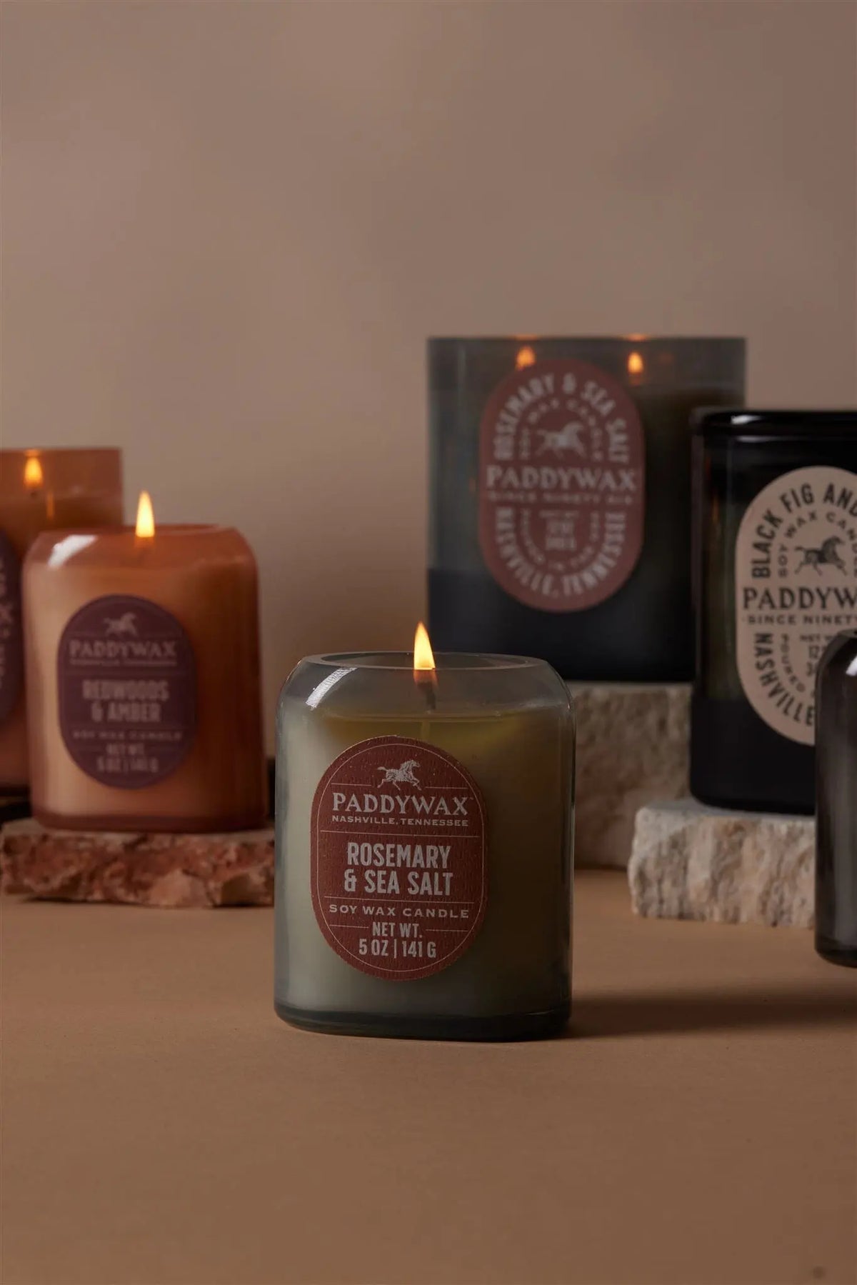 A lit Rosemary + Sea Salt - Vista 12 oz Candle from the vintage-inspired collection is displayed on a neutral surface with stone blocks.