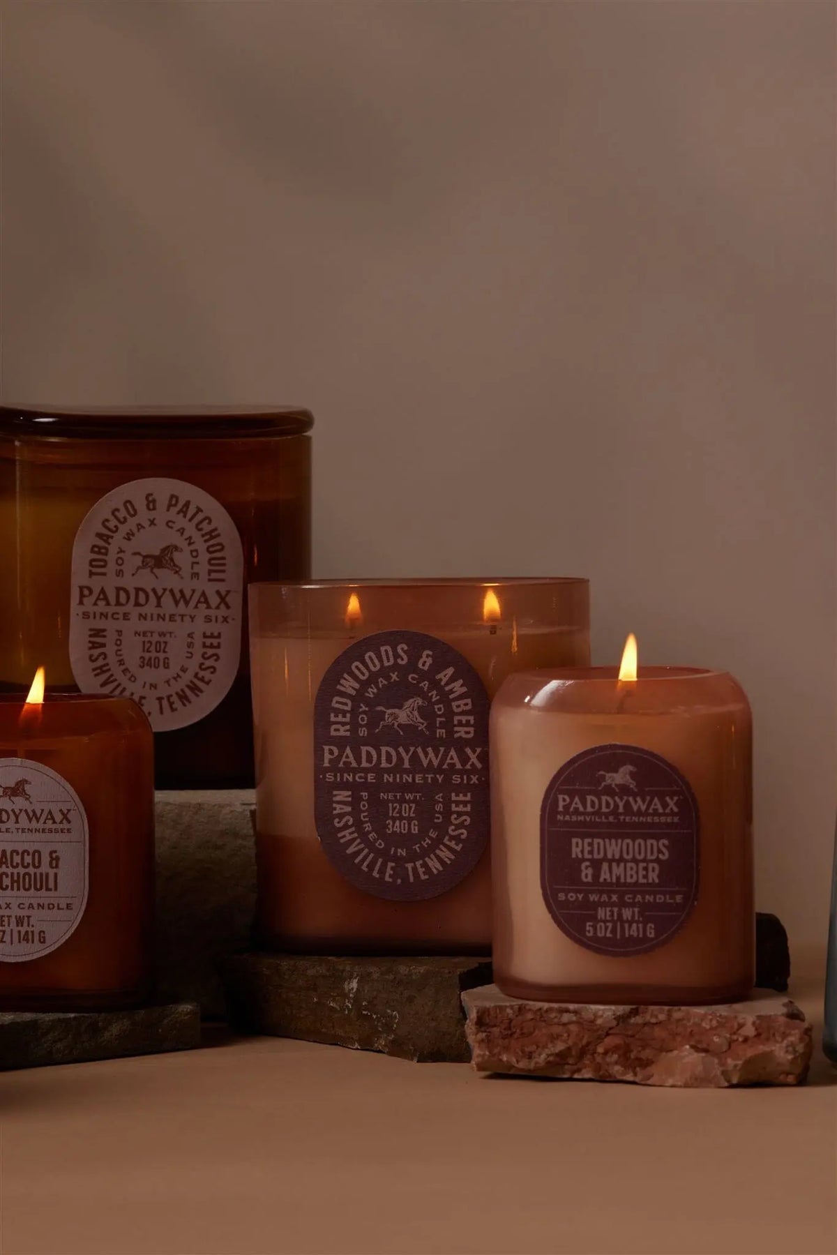 Four vintage-inspired Vista 12 oz candles, including the Redwoods + Amber scent, are each housed in elegant amber and beige glass vessels and artfully arranged on stacked stone slabs against a neutral background.