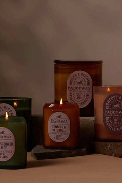 Five lit Tobacco + Patchouli - Vista 5 oz Candles by Paddywax in amber and green glass jars are arranged on stone slabs, bringing modern decor flair as their labels highlight the luxurious fragrance.