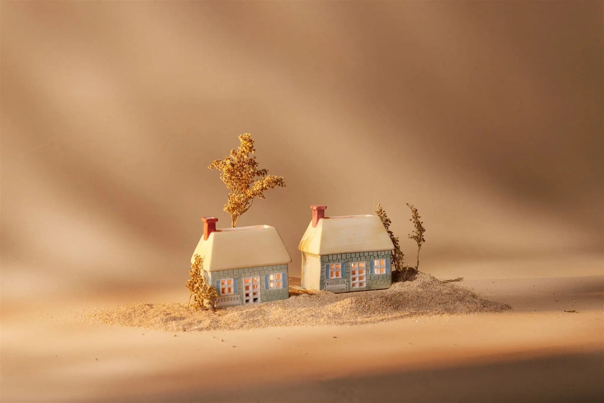 The No. 04 The English Cottage Incense & Tea Light Holders feature two small ceramic houses with lit windows on sand, surrounded by dried plants—a charming decor piece for creating a cozy cottage atmosphere.