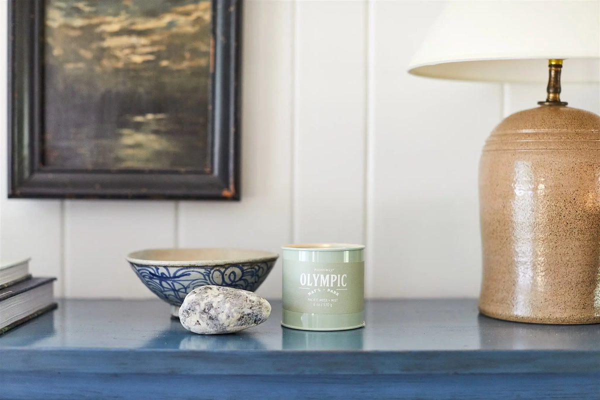 An Olympic - Parks 6 oz Tin Candle, ceramic lamp, patterned bowl, and two stones are arranged on a blue table with a framed painting in the background.