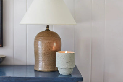 A ceramic table lamp with a white shade is on a blue surface beside a lit Glacier - Parks 11 oz Candle in a speckled cup-shaped holder with a crackling wood wick.