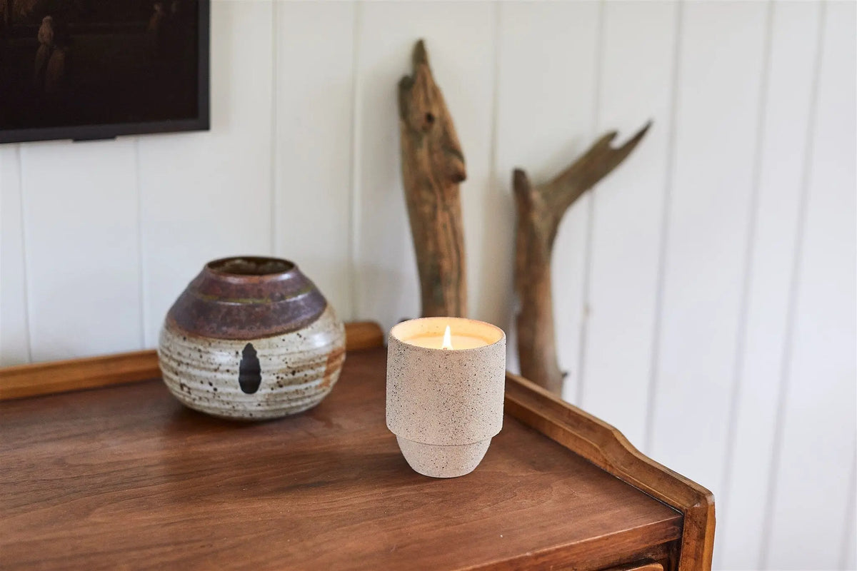 The Yellowstone - Parks 11 oz Candle with a crackling wood wick glows in a ceramic holder on a wooden table beside a pottery vase and driftwood, all set against a white paneled wall.