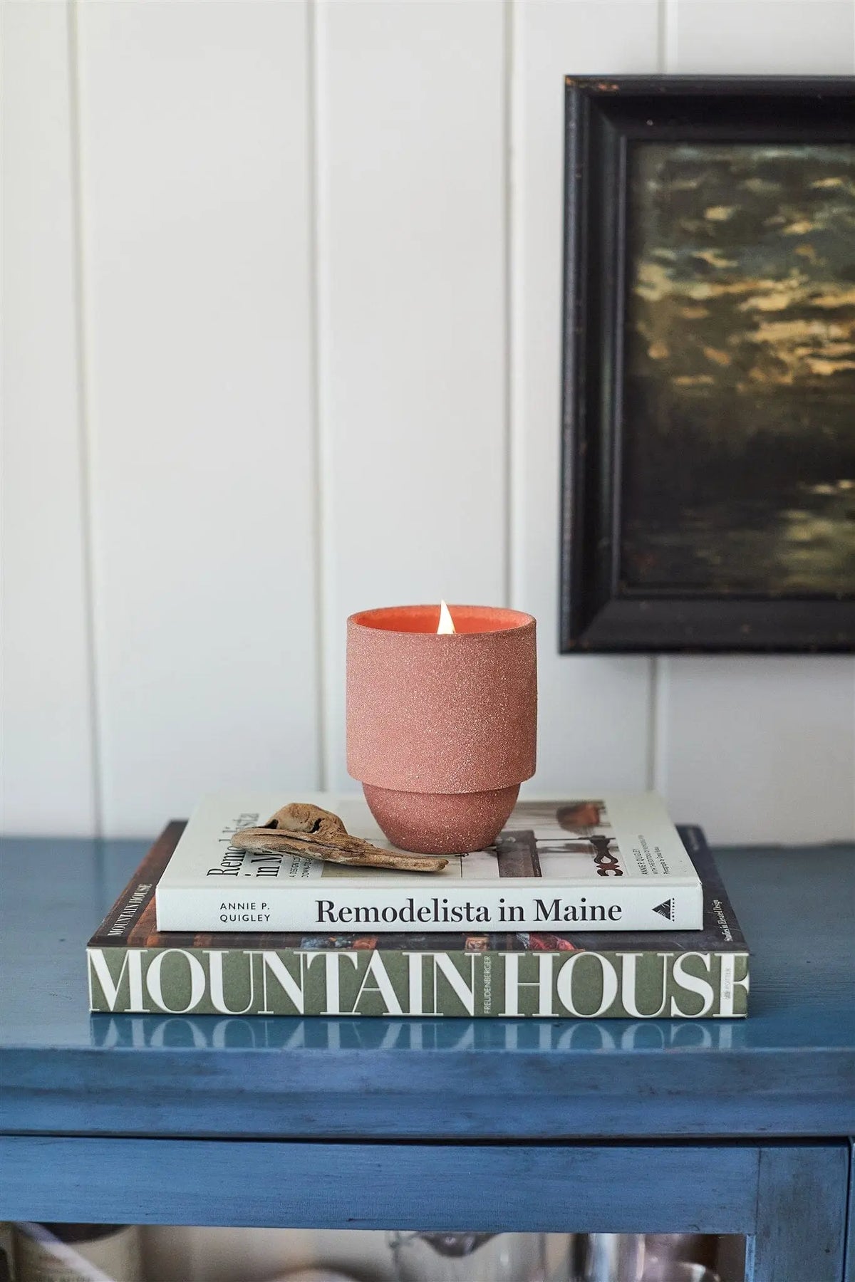 The Redwoods - Parks 11 oz Candle, featuring a crackling wood wick and pink wax, sits atop two books on a blue table. Its woody Redwood and Lily scent is inspired by Redwoods National Park. A framed painting appears in the background.
