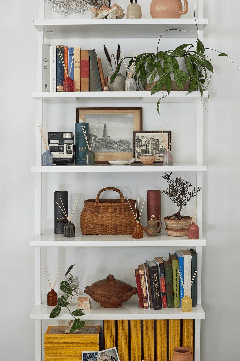 A white shelving unit with five shelves displays books, framed photos, a plant, vases, a wicker basket, and decorative objects—including the Fresh Air 1.5 oz Diffuser—artfully arranged for a stylish look.