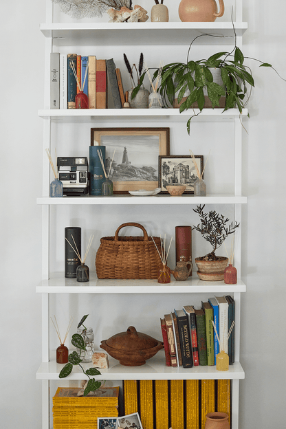 A white shelving unit with five shelves displays books, framed photos, a plant, vases, a wicker basket, and decorative objects—including the Fresh Air 1.5 oz Diffuser—artfully arranged for a stylish look.