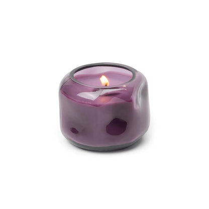 A small, round, purple glass candle holder houses the Violet & Vanilla - Basalt 5 oz Candle, its warm flame casting a gentle glow and filling the air with violet and vanilla scents.