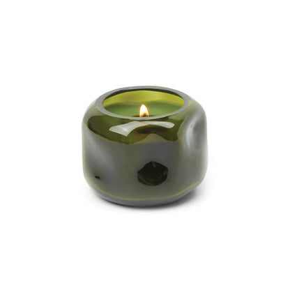 A compact, round green glass vessel houses the Wild Fig & Cedar - Basalt 5 oz Candle, its gentle flame illuminating the earthy-woody scent against a simple white background.