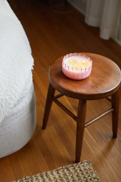 A Desert Peach - 12 oz Candle in vintage glass with multiple wicks sits on a wooden stool by a bed with a white blanket, on hardwood flooring.