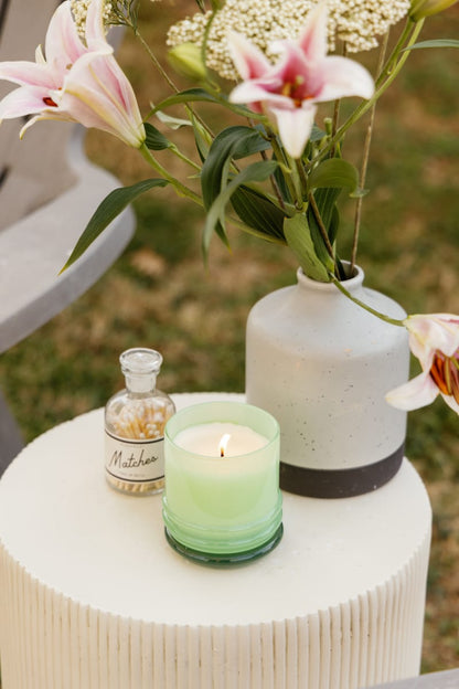 A small white table on grass holds a lit Minty Verde - Stack 7 oz Candle in a reusable vessel, a glass bottle labeled "Matches," and a ceramic vase with pink lilies and white flowers, all set outdoors.