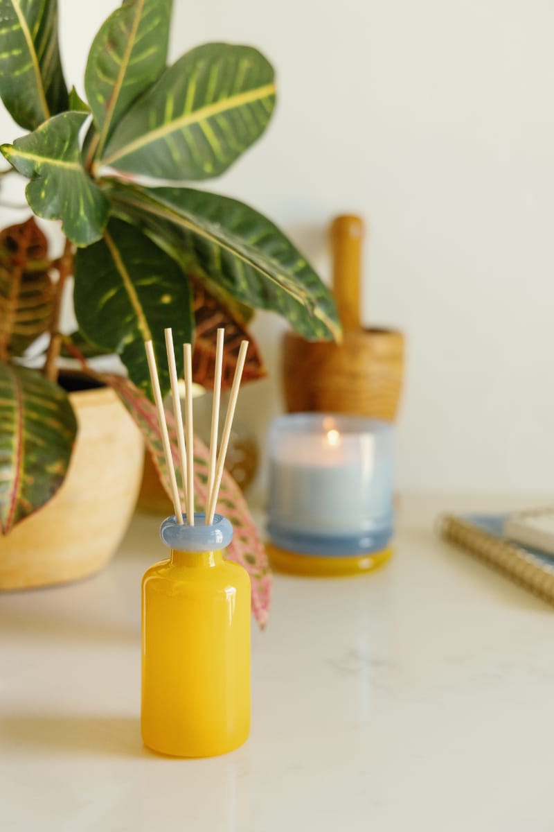 A Zesty Lemon - Stack 3.2 oz Diffuser with wooden sticks sits on a white surface, surrounded by a potted plant, a lit candle in a blue holder, and a wooden mortar and pestle for a modern home fragrance vibe.
