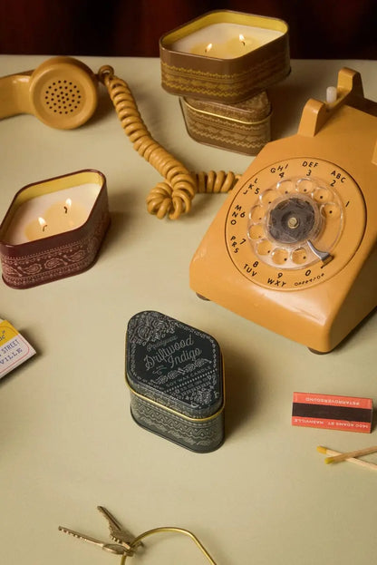 On a pale surface, a vintage yellow rotary phone is displayed alongside woody Driftwood Indigo - Station Tin 3.5 oz candles inspired by National Parks scents in travel-sized patterned tins, matches, keys, and a phone receiver.