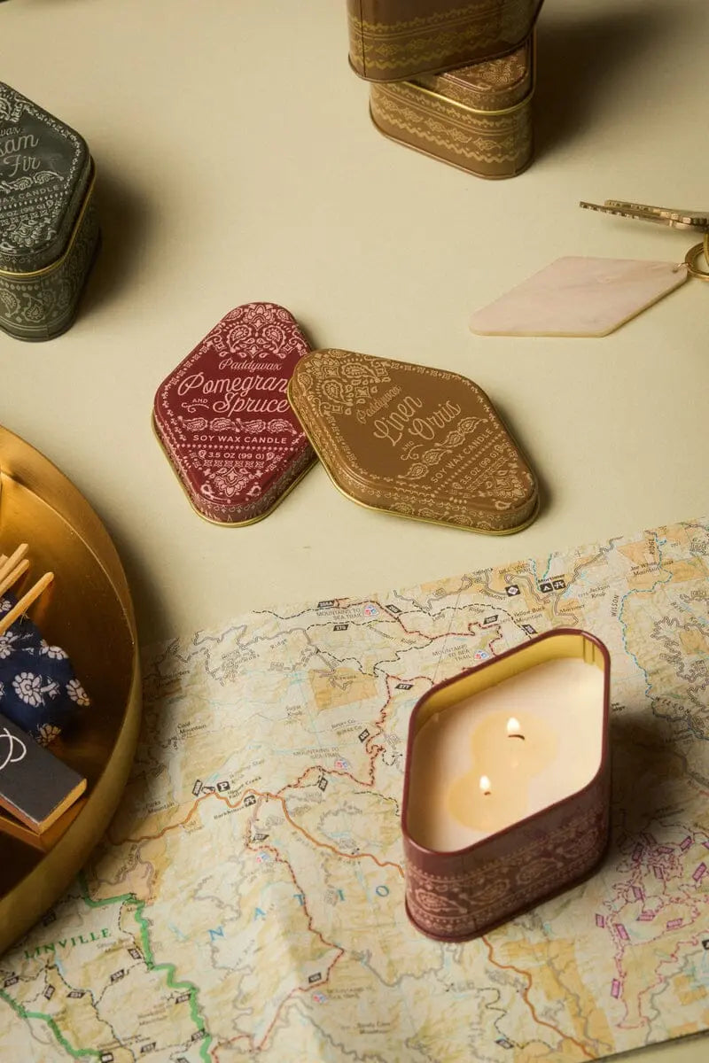 A 3.5 oz Station Tin Candle in the Pomegranate Spruce scent—with notes of woody freshness inspired by Northern California—sits atop a map in a decorative, travel-friendly patterned tin. Nearby are closed tins, including Linen & Paris, evocative of airy and refined fragrances. Also visible are a gold tray with matchboxes and a marble-handled letter opener.