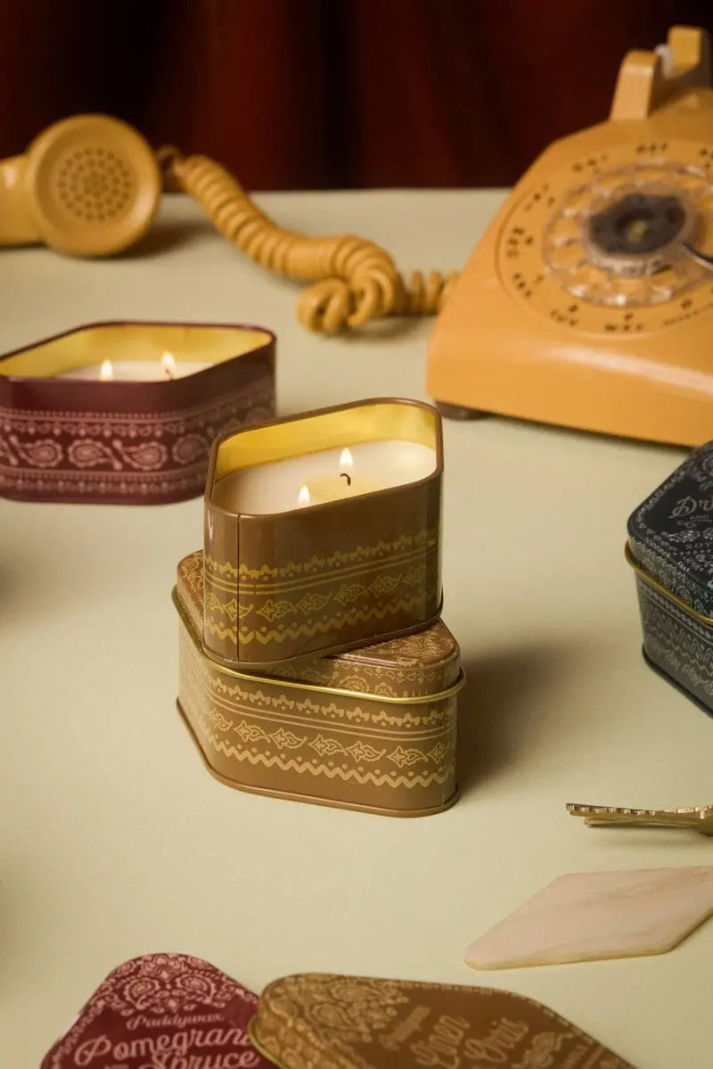 A Linen & Orris - Station Tin 3.5oz candle sits lit on a table among other tins, filling the air with its signature woody notes inspired by Northern California, while an old rotary phone and coiled cord lend vintage charm in the background.