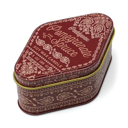 Decorative maroon tin in a rounded triangle shape featuring ornate cream lettering and floral patterns on the lid, labeled “Pomegranate Spruce - Station 3.5 oz Tin Candle,” inspired by the woody scents of Northern California.