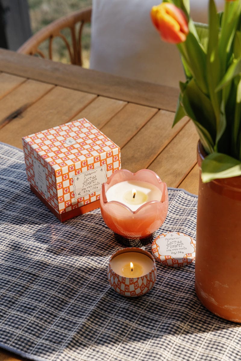 A wooden table displays a potted yellow and orange tulip in a peach vessel, two lit Cactus Flower - Tulip 5.5 oz Candles in patterned containers, and a matching box, all atop a checkered table runner.