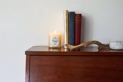 A Worn Leather & Smoke - Vista 5 oz Candle in a glass jar rests on wood beside three upright books, driftwood, and a smooth gray stone, creating a calm modern decor scene against a plain white wall.