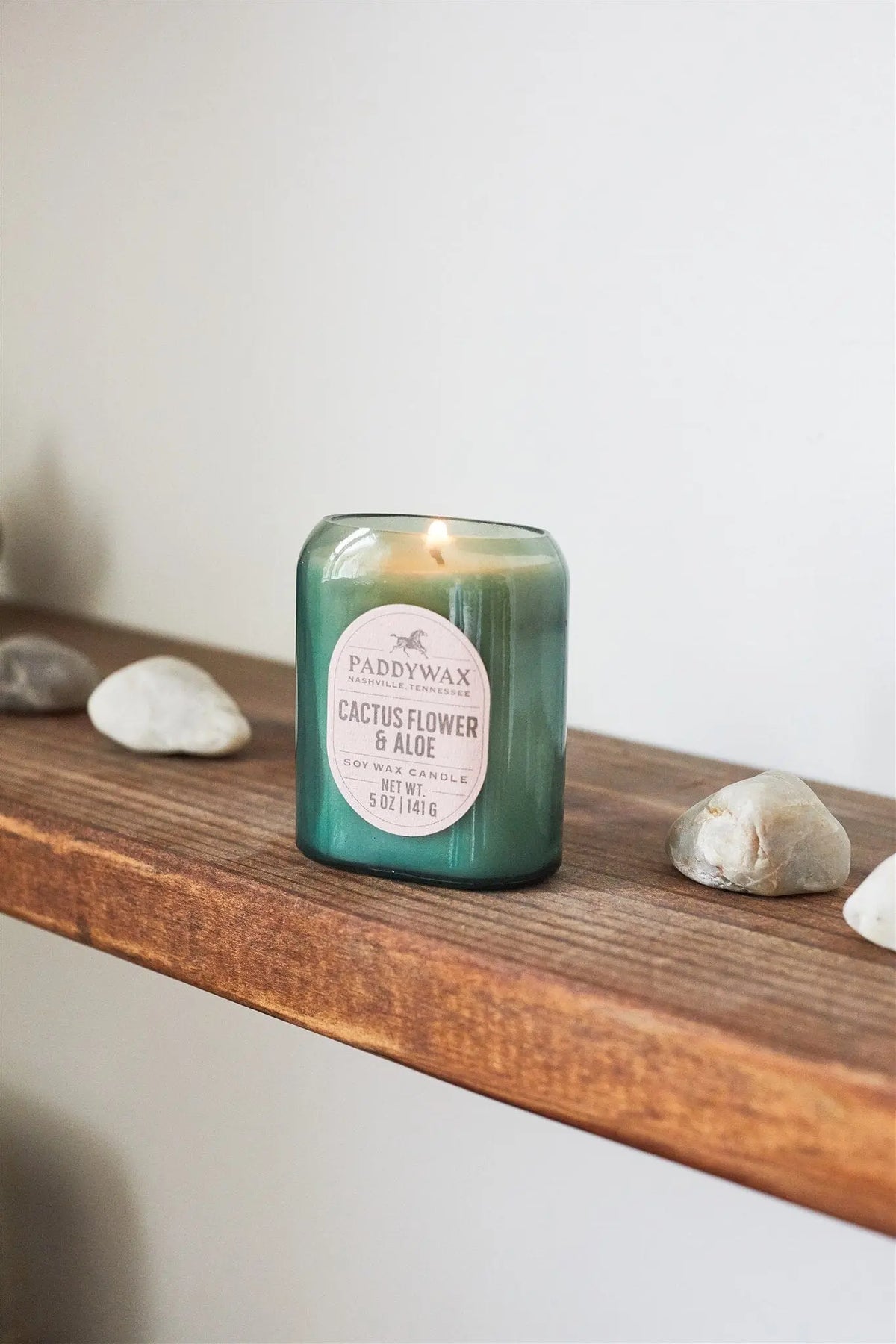 A Cactus Flower & Aloe - Vista 5 oz Candle burns on a wooden shelf, its luxurious fragrance enhancing modern decor while smooth white and beige stones surround it.