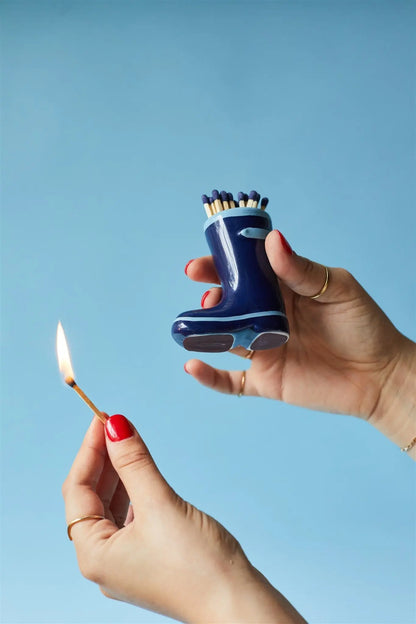 A person holds the Wellington Boot Matches - Navy Blue, filled with safety matches, and lights one matchstick with the other hand against a blue background.