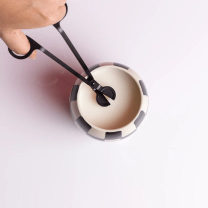 A hand uses the Wick Trimmer - Matte Black to trim the wick of a candle in a black-and-white striped container on a white surface.