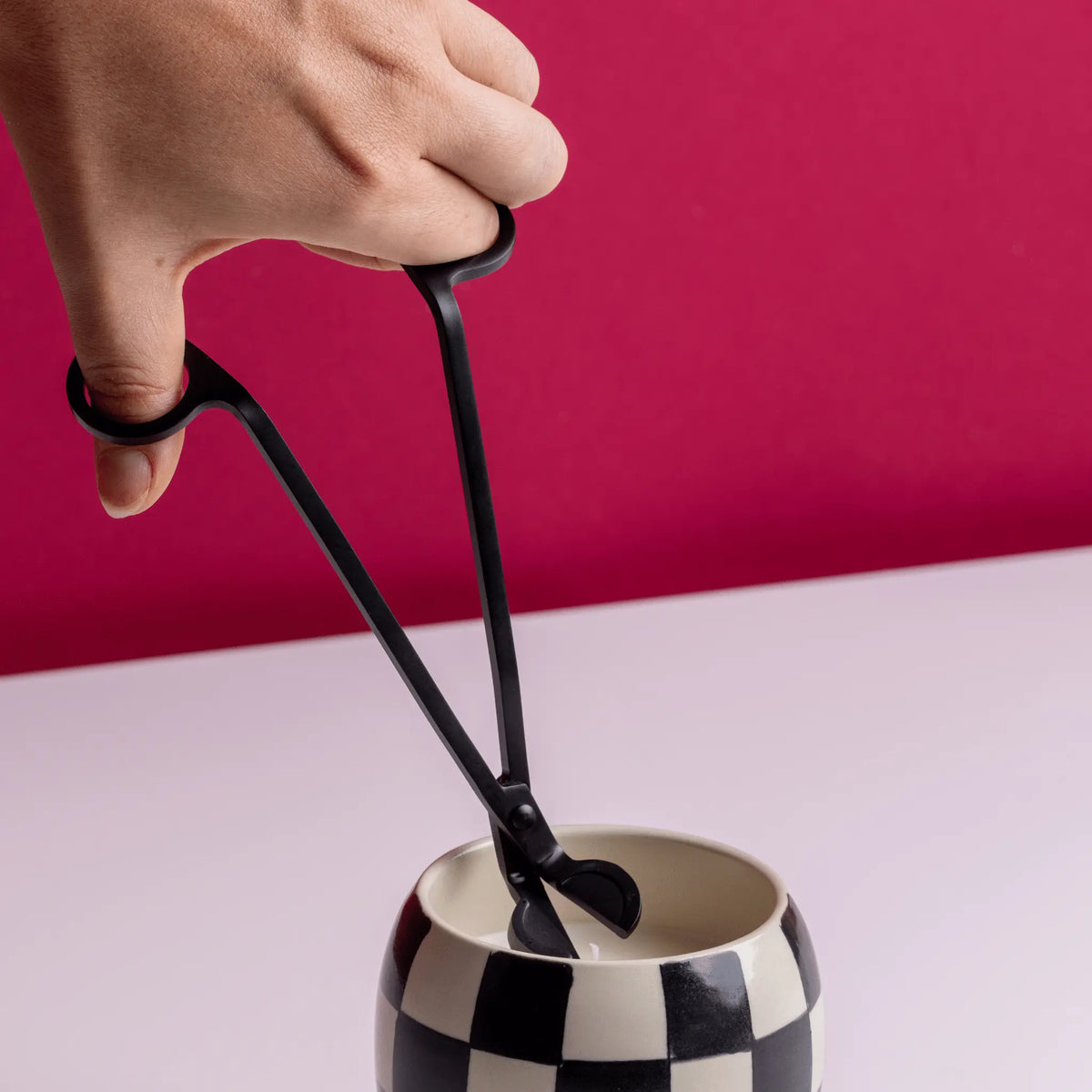 A hand uses the Wick Trimmer - Matte Black to neatly trim the wick of a candle in a black-and-white checkered holder, set against a pink background.