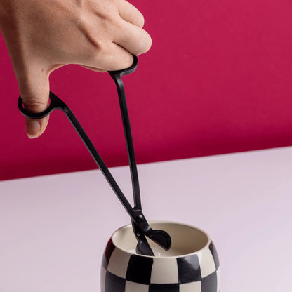 A hand uses the Wick Trimmer - Matte Black to neatly trim the wick of a candle in a black-and-white checkered holder, set against a pink background.