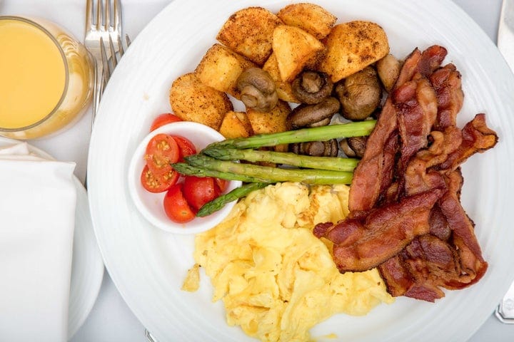 A plate with scrambled eggs, crispy bacon strips, roasted potatoes, sautéed mushrooms, cherry tomatoes, and asparagus, served with a glass of orange juice and cutlery on a white tablecloth.