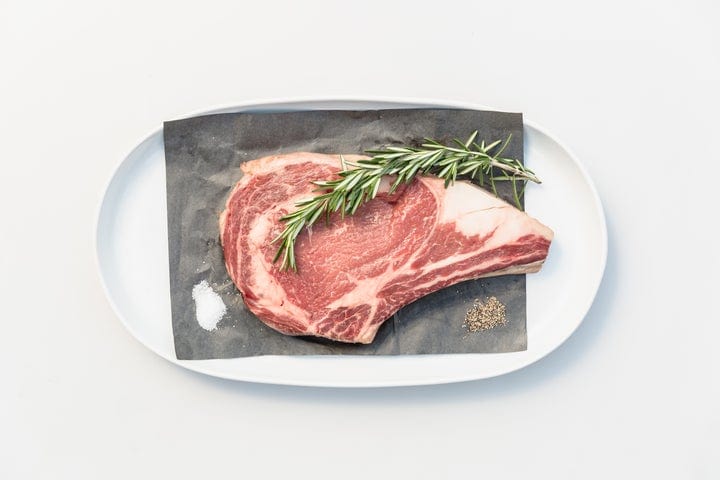 A raw ribeye steak with a sprig of rosemary rests on black parchment paper, accompanied by small piles of salt and pepper, all presented on a white oval plate against a white background.