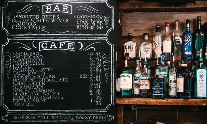 Chalkboard menu displaying bar and café drink options with prices. Bottles of liquor are arranged on shelves beside the menu, which lists beers, wines, coffee drinks, and other beverages.