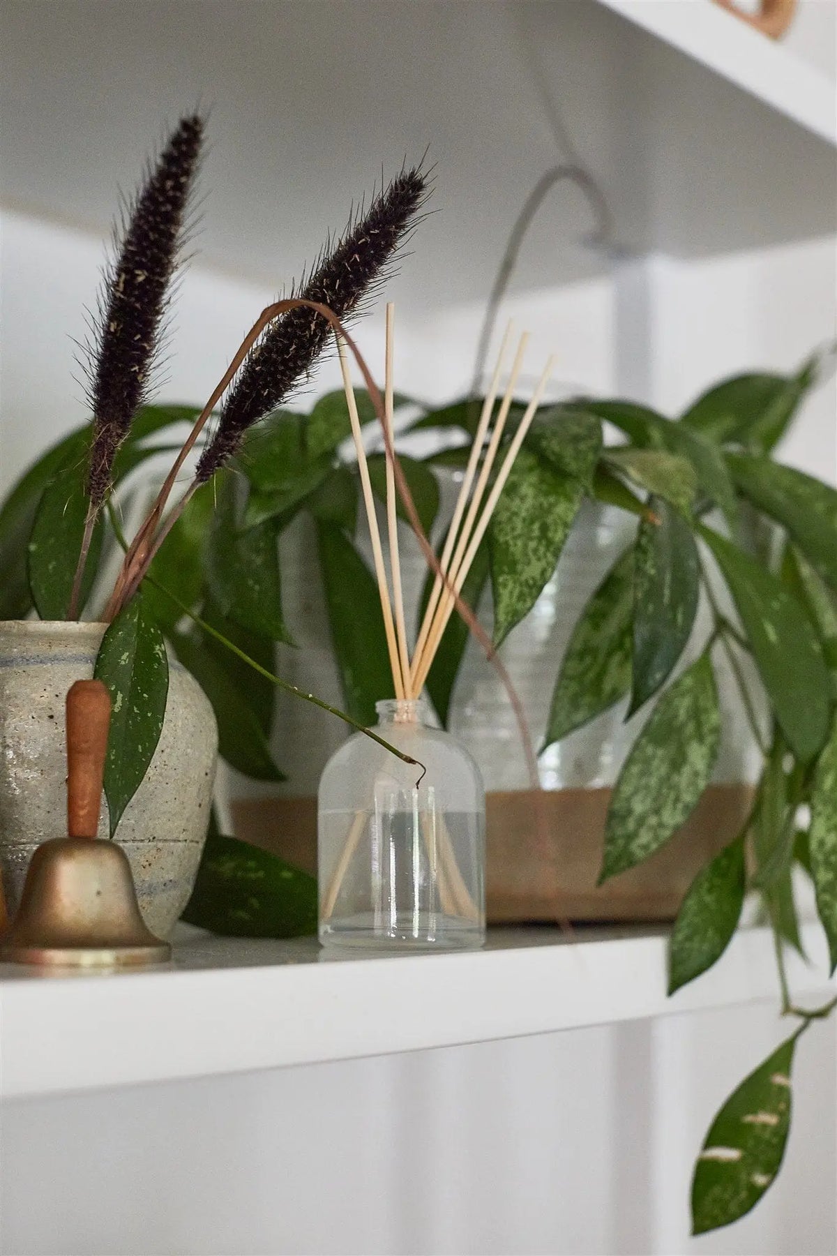 A white shelf displays a potted plant, a small brass bell, a ceramic vase with dried grass, and the Flowers - 1.5 oz Diffuser—a milky glass fragrance gift with wooden reeds for the perfect presentation.