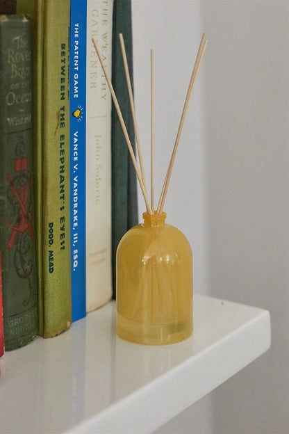 A Lemon - 1.5 oz Diffuser with wooden reeds sits on a white shelf next to upright books with colorful spines.