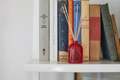 A close-up of a white bookshelf with several books upright and the Cranberry - 1.5 oz Diffuser in front, adding a touch of elegance and subtle fragrance to the scene.